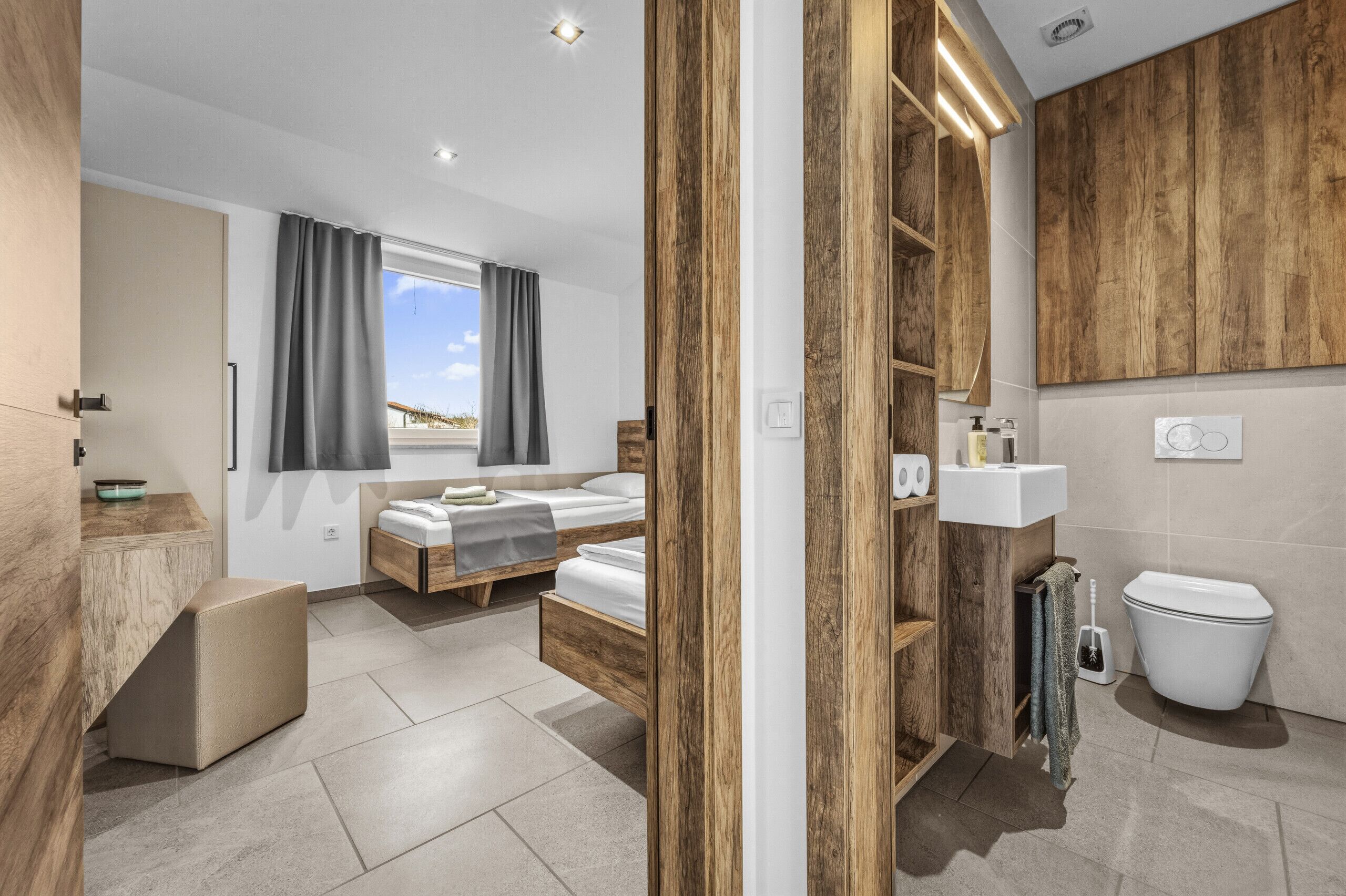 A modern bedroom with a large window, a wooden wardrobe, a bedside table, and a bathroom with a toilet and sink.
