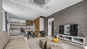A modern living room with a sofa, ottoman, and a TV on a white stand. There is a kitchen area with cabinets and a dining table. - Lavanda Harmony Apartment (Pomer)