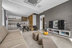 Ein modernes Wohnzimmer mit einer Couch, einem Hocker und einem Fernseher auf einem weißen Schrank. Es gibt eine Küchenzeile mit Schränken und einem Esstisch.