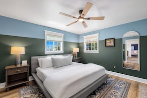 A bedroom with a bed, nightstands, a ceiling fan, two windows with blinds, a mirror, and a patterned rug.