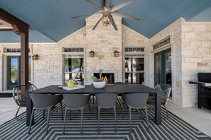 An outdoor patio with a stone wall, a ceiling fan, a table with chairs, and a fireplace.