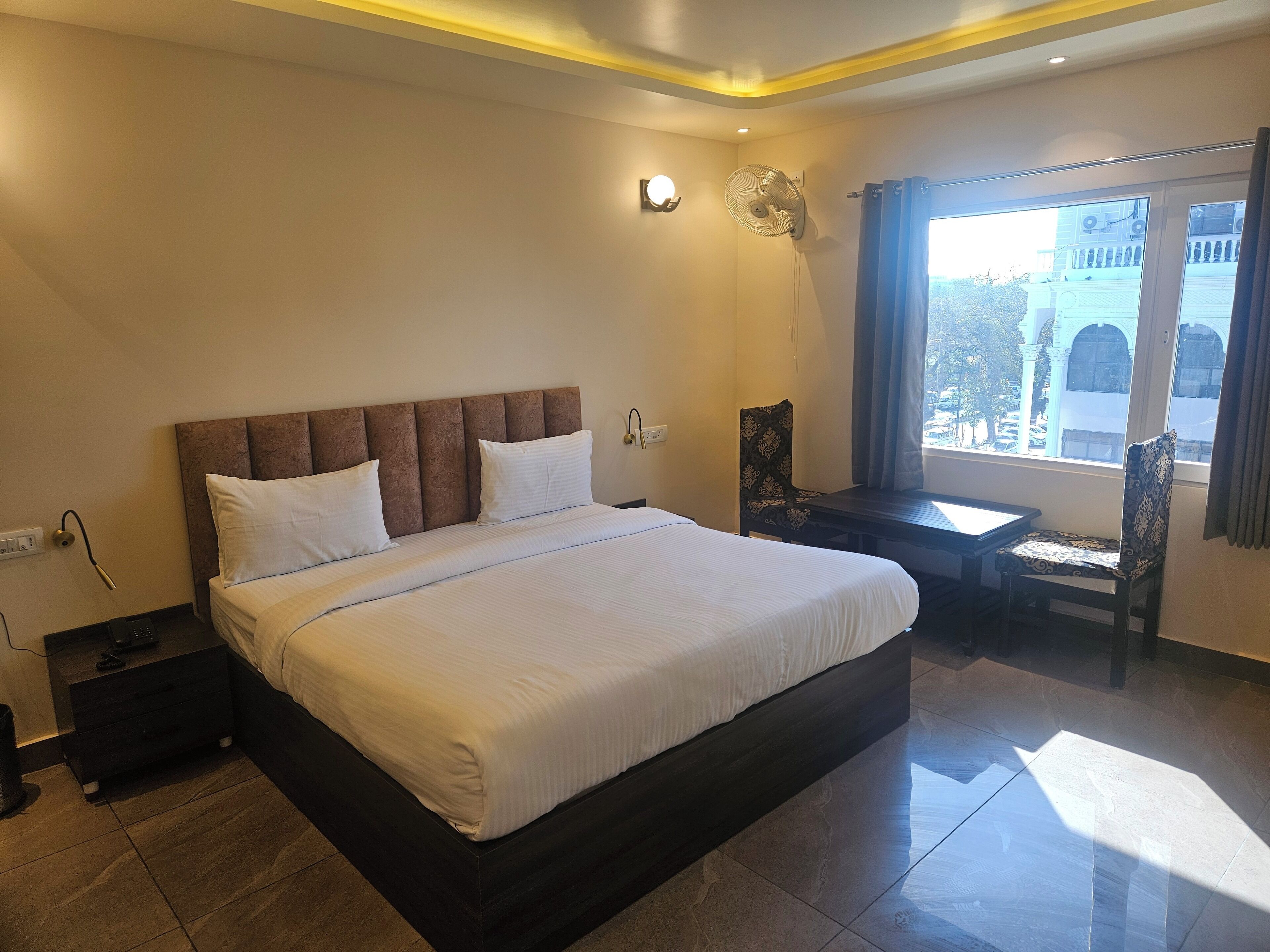 A hotel room with a large bed, a desk, and a chair. There is a window with curtains, a wall-mounted fan, and a nightstand with a lamp.