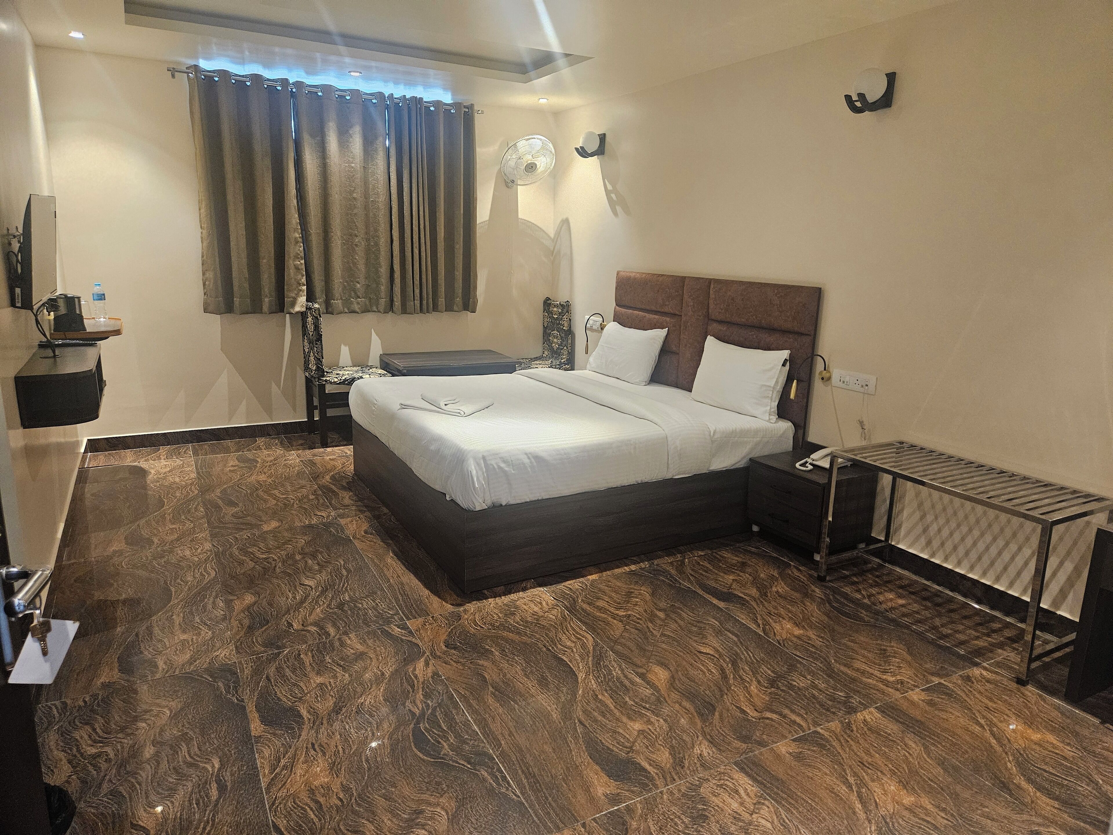 Una habitación de hotel ordenada con una cama grande, una mesita de noche, un escritorio, una silla, un ventilador de pie y un radiador.