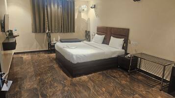 Una habitación de hotel ordenada con una cama grande, una mesita de noche, un escritorio, una silla, un ventilador de pie y un radiador.
