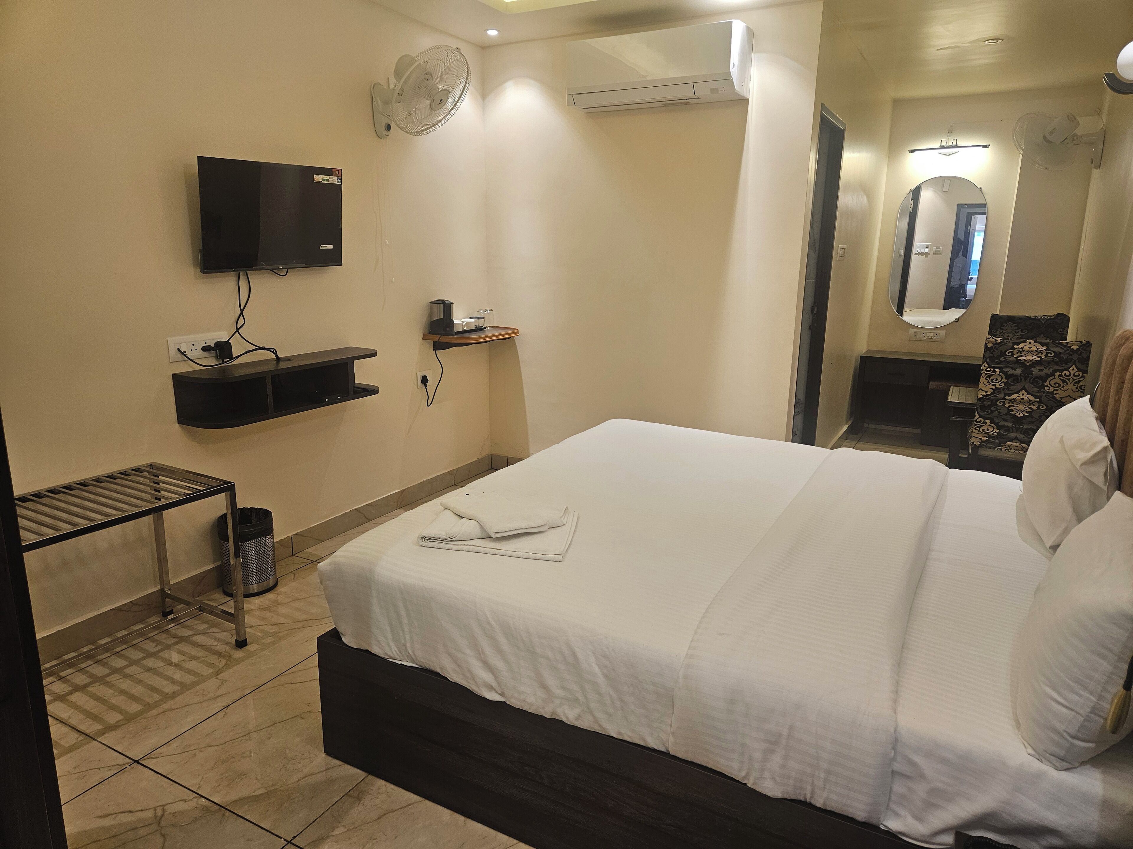 Una habitación de hotel con cama, televisor, aire acondicionado, espejo, una mesita con una taza y un vaso, y un cubo de basura.