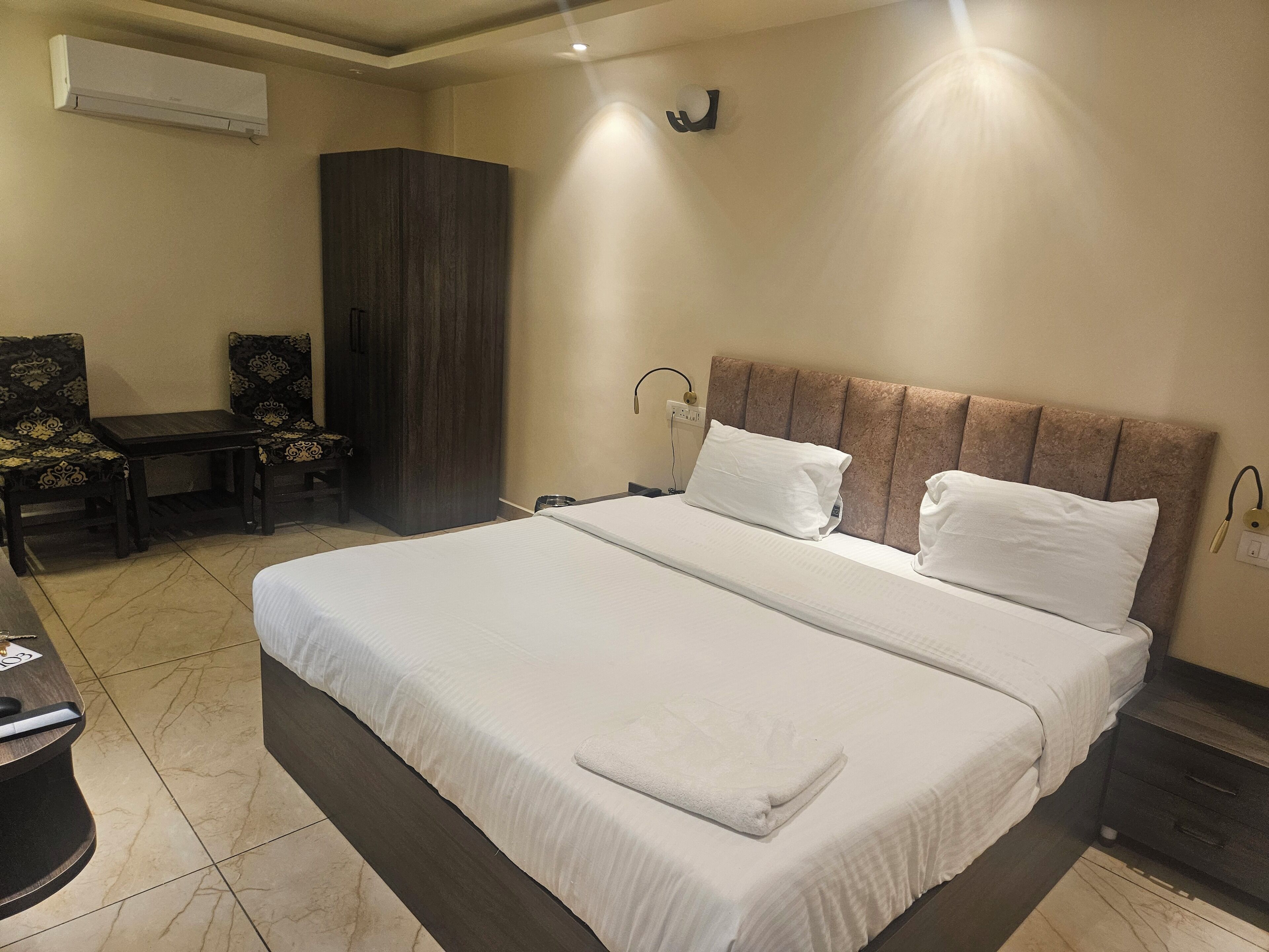 Une chambre d’hôtel avec un grand lit, un bureau, deux chaises, une armoire et un climatiseur fixé au mur.