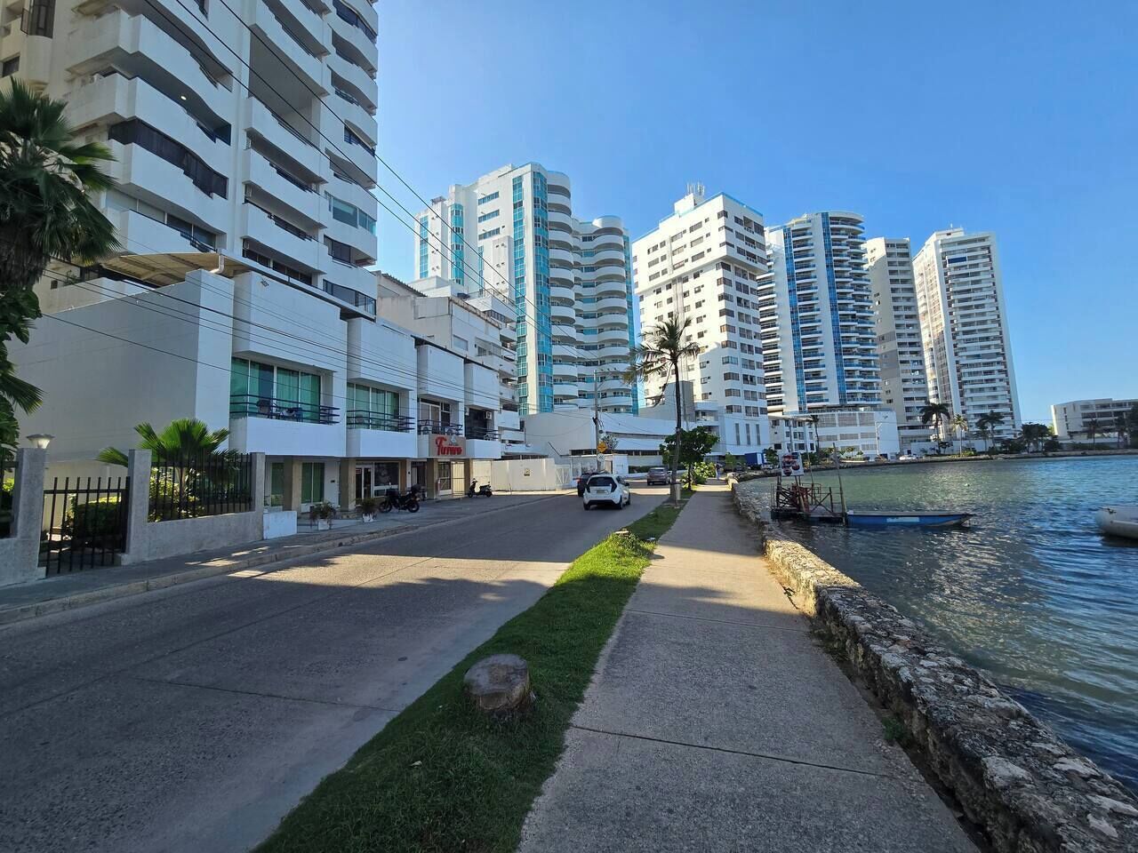 A coastal urban street with modern high-rise buildings, a sidewalk, and a body of water.