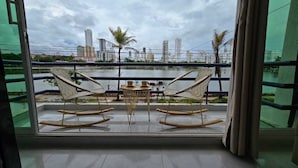 Een balkon met twee schommelstoelen en een kleine tafel, met uitzicht op een waterpartij en een stadsgezicht met hoge gebouwen.