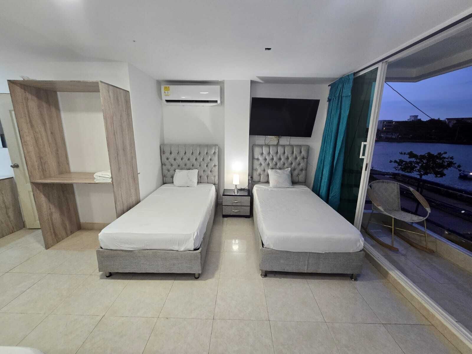 A hotel room with two beds, a TV, a window with a view of the outside, and a small table with a lamp.