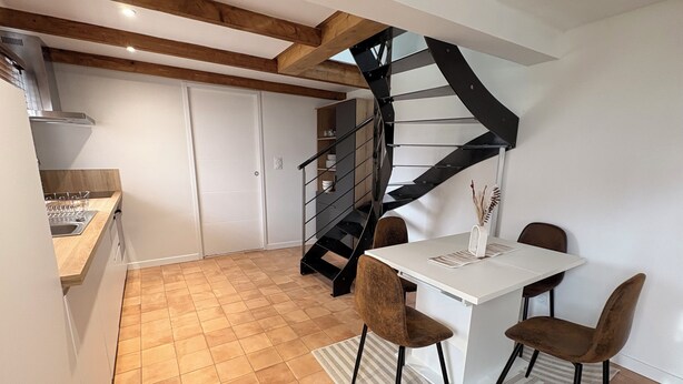 Una cocina moderna con horno empotrado, una zona de comedor con mesa blanca y sillas marrones, y una escalera espiral negra que conduce a una habitación superior.