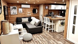 A cozy cabin interior with a wooden ceiling, a sofa with cushions, a dining area with chairs, a kitchen with white cabinets, and a small round table.