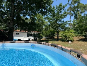 A swimming pool with a wooden deck, surrounded by trees and a white RV in the background.