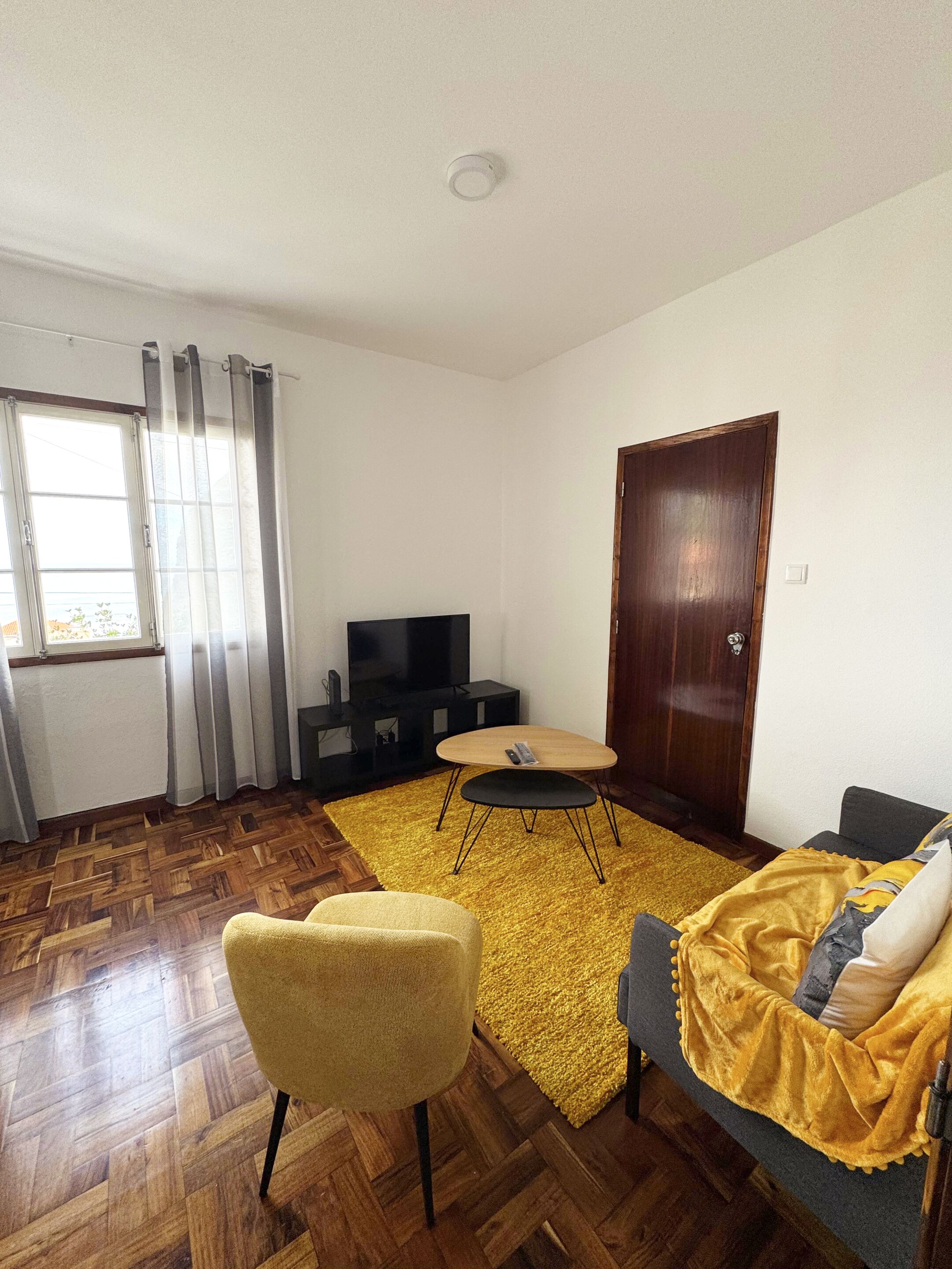 Uma sala de estar com piso de madeira em padrão herringbone, um tapete amarelo, uma poltrona amarela, um sofá com manta amarela, uma porta de madeira e uma televisão sobre um móvel preto.