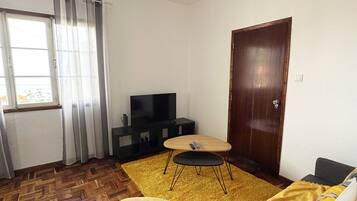 Uma sala de estar com piso de madeira em padrão herringbone, um tapete amarelo, uma poltrona amarela, um sofá com manta amarela, uma porta de madeira e uma televisão sobre um móvel preto.