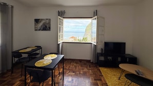 A compact living space with a dining area, a TV, and a window with a view of the sea. - Ocean View Guest House (Sao Vicente)