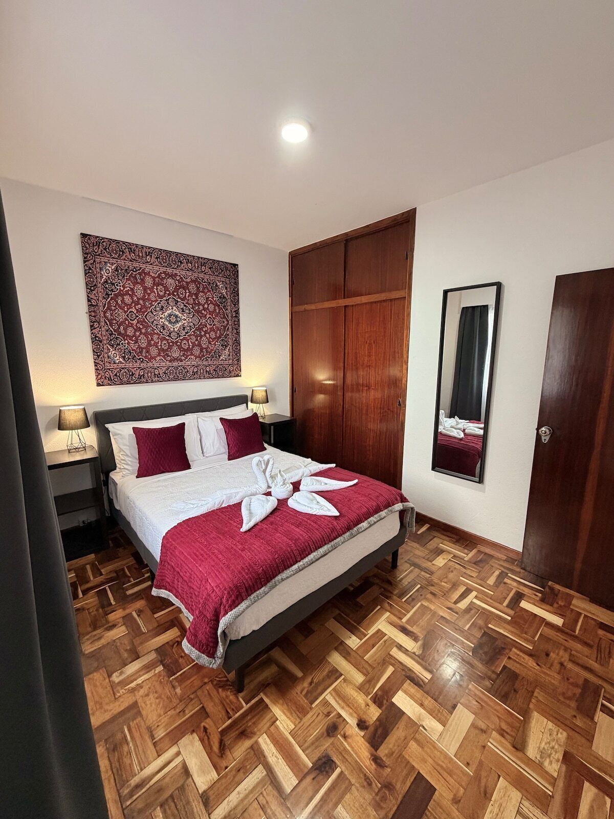 A hotel room with a wooden herringbone floor, a bed with a red and white bedspread, a large mirror, and a wooden wardrobe.