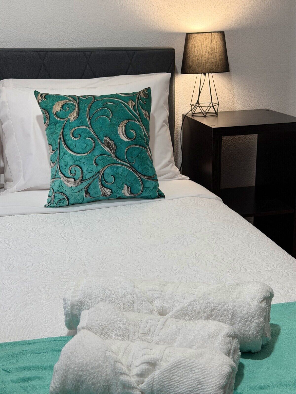 A neatly made bed with a teal patterned pillow, a white blanket, and two white towels on a green bedspread.