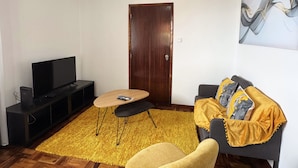 A living room with a wooden herringbone floor, a yellow armchair, a dark sofa with a yellow throw, a wooden coffee table, a flat-screen TV on a black stand, and a wooden door. - Ocean View Guest House (Sao Vicente)