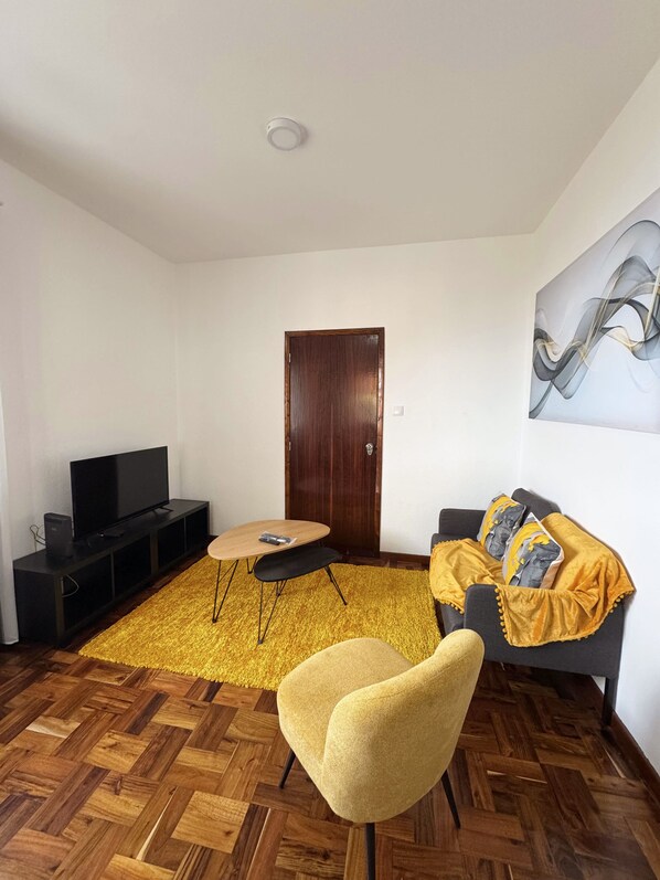 A living room with a wooden herringbone floor, a yellow armchair, a dark sofa with a yellow throw, a wooden coffee table, a flat-screen TV on a black stand, and a wooden door.