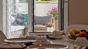 En matplats med ett bord dukat för en måltid, en soffa i rotting med kuddar och en utsikt över utomhusmiljön genom en glasdörr.