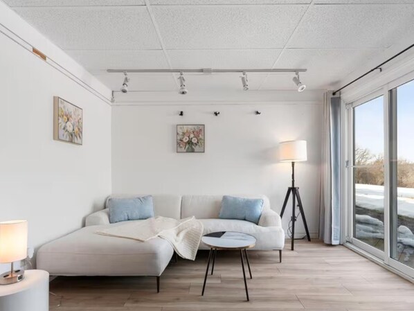 Un salón moderno con un sofá blanco, una mesita redonda y una lámpara de pie. Hay luces de techo y un amplio ventanal con vistas al exterior.