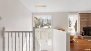 A room with a white door, a brick fireplace, and a wooden staircase. - Large Whole Home in Wine Country: Pool Hottub Sauna + Spacious Backyard & Nature (Prince Edward)