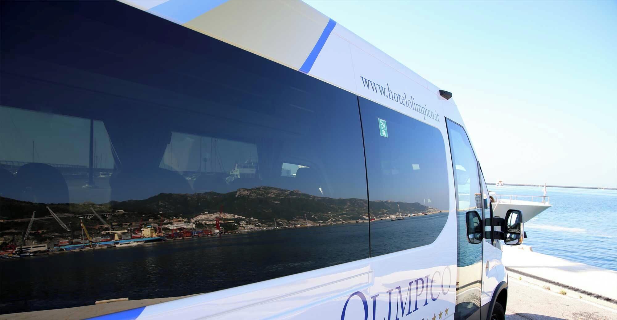 Un autobus di grandi dimensioni con il sito web 'www.hotelolimpico.it' sul fianco, parcheggiato vicino all'acqua, con vista su un porto e un ponte sullo sfondo.