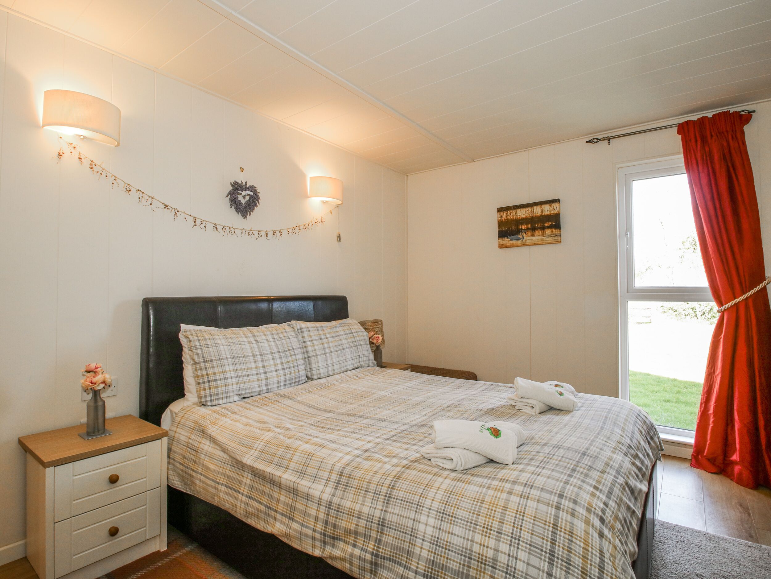 Une chambre à coucher comprenant un lit, des tables de chevet, une commode, une lampe fixée au mur, une fenêtre avec des rideaux et un petit élément décoratif mural.