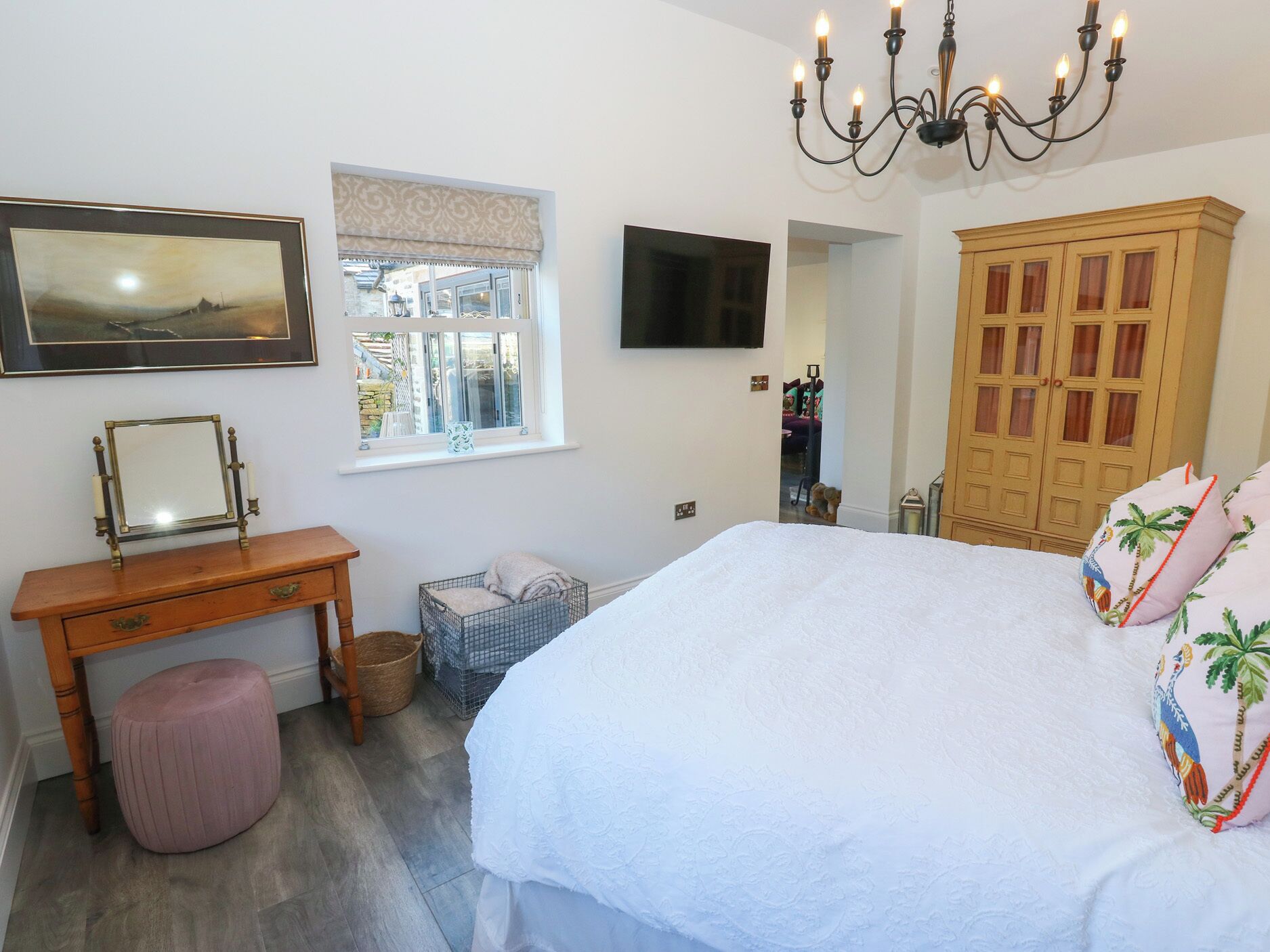 A bedroom with a bed, a wooden dresser, a vanity with a mirror, a television, a window with a view, and a chandelier.