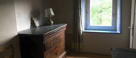 A room with a wooden dresser, a lamp, a window with curtains, and a chair.