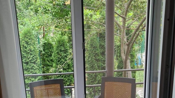 A room with a large window overlooking a lush green area with trees and a building in the background. - Cozy Corner (Borjomi)
