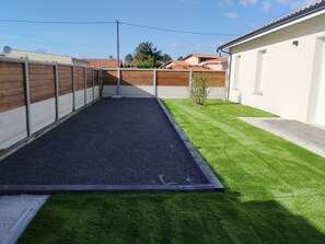 A fenced backyard with artificial grass, a gravel area, and a white building with a tiled roof. - New house with swimming pool. Family vacation (Gujan-Mestras)