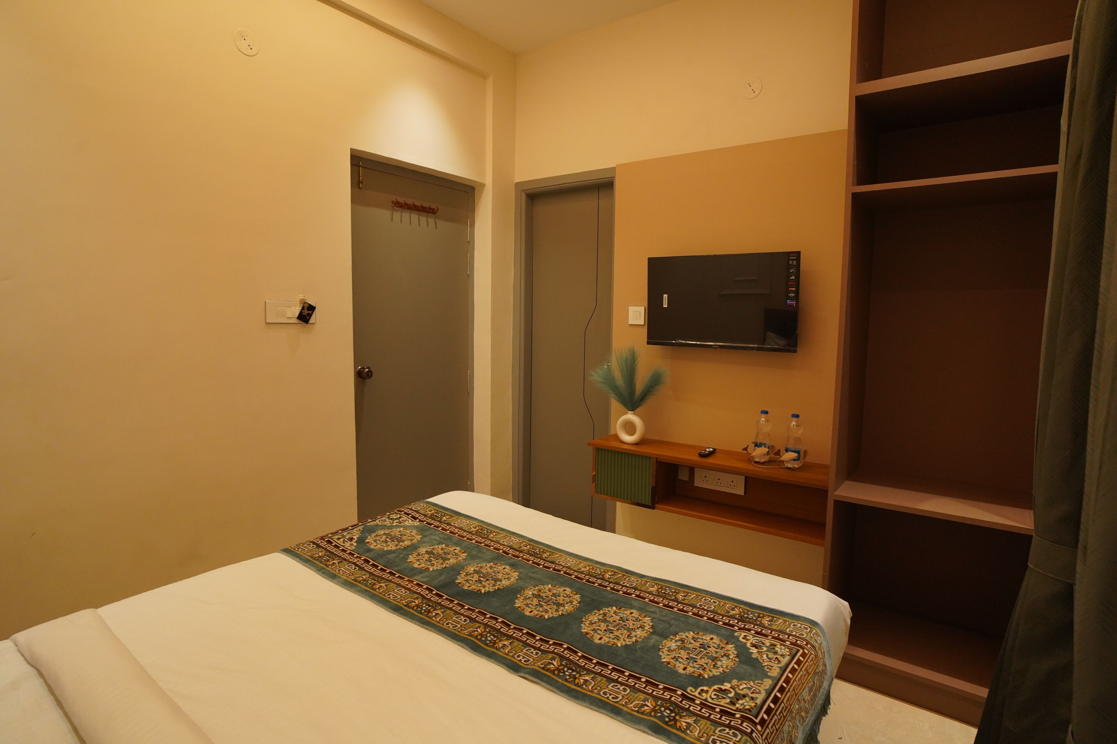 A hotel room with a bed, a television mounted on the wall, a small shelf with bottles, and a wardrobe.