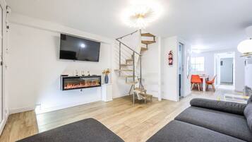 A modern living room with a fireplace, a flat-screen TV, a staircase, and a dining area with orange chairs.