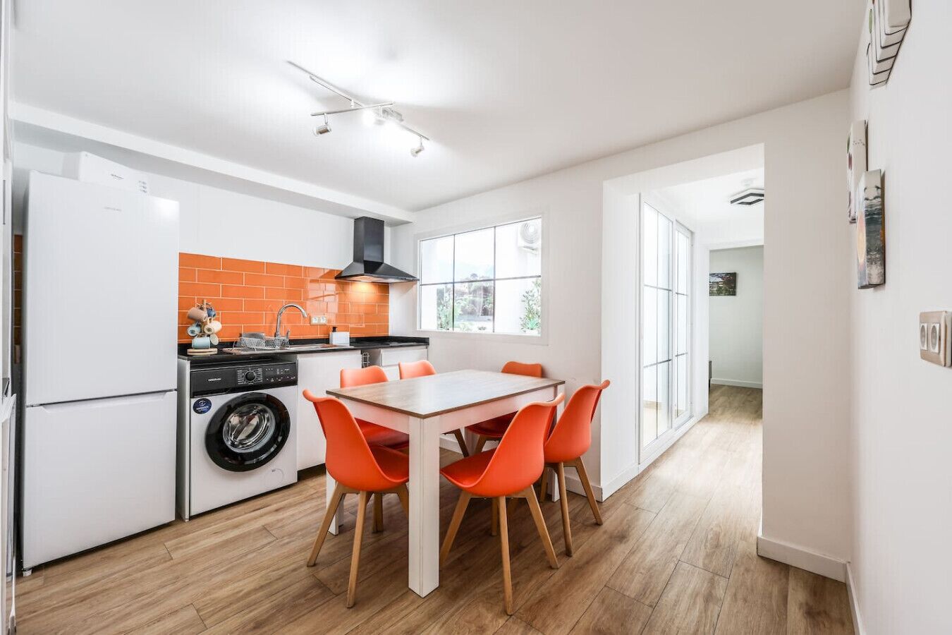 A modern kitchen with a washing machine, a dining table with orange chairs, and a large window providing natural light.