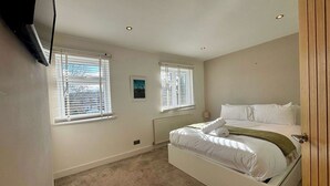 A bedroom with a bed, two windows, a radiator, and a television mounted on the wall. - NEC Solihull Home | Parking, Garden, JLR & Airport (Solihull)