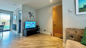 A modern living room with a flat-screen TV, wooden flooring, a brown leather sofa, and a sliding glass door leading to a patio. - NEC Solihull Home | Parking, Garden, JLR & Airport (Solihull)