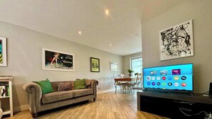 A living room with a wooden floor, a grey sofa, a TV, and framed pictures on the wall. - NEC Solihull Home | Parking, Garden, JLR & Airport (Solihull)