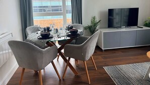 Ett modernt vardagsrum med ett matbord för fyra personer, en grå soffa, en tv-apparat på ett skåp och utsikt över en byggnad genom stora fönster.