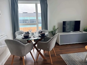 A modern living room with a dining table set for four, a grey sofa, a TV on a cabinet, and a view of a building through large windows. - 2BR Bracknell Apt Near Ascot | Parking & WiFi (Bracknell)