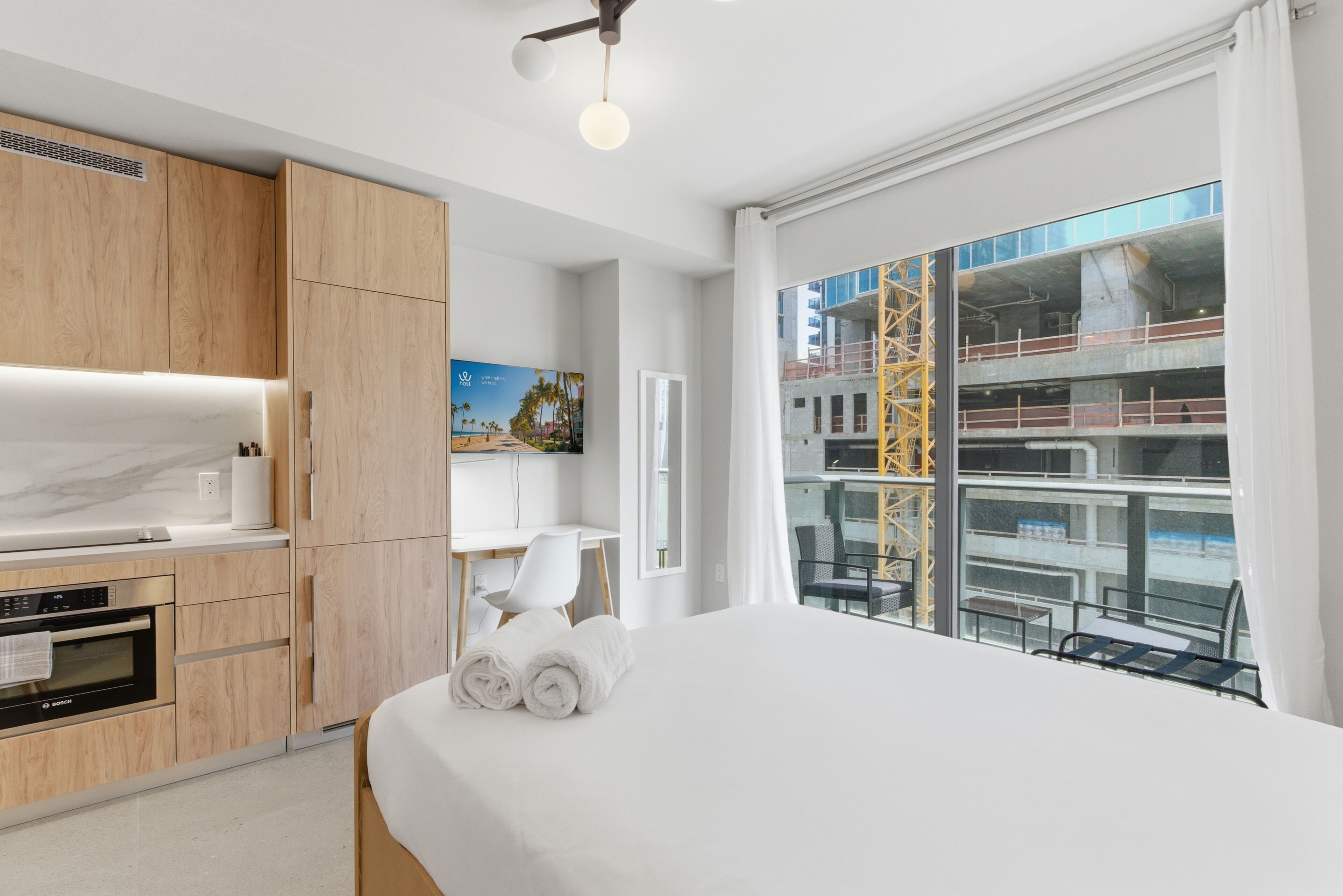 A modern hotel room with a bed, a kitchen area, a dining table, and a balcony with a view of the city.
