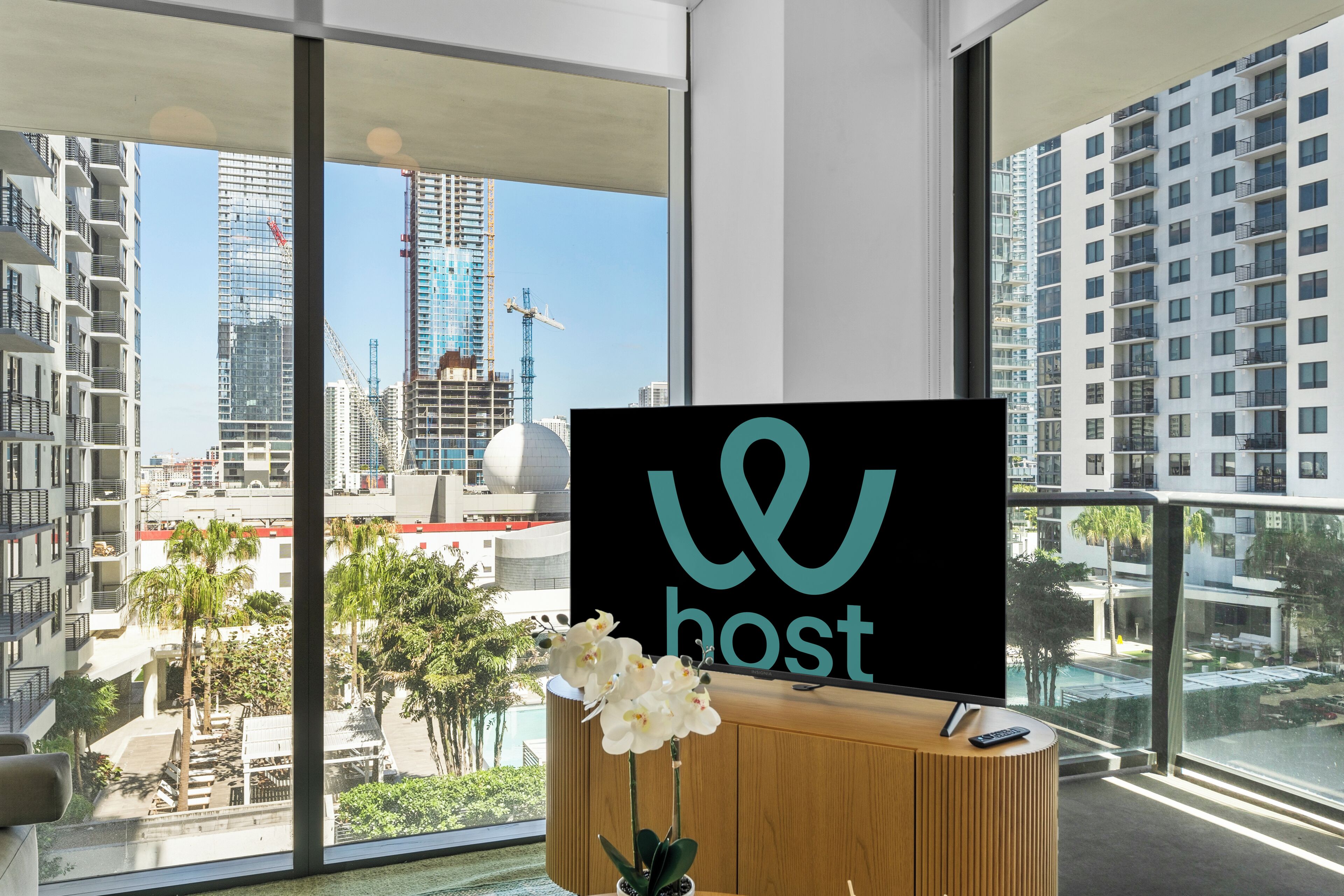 Ein modernes Hotelzimmer mit großem Fenster und Blick auf die Stadt, ein Holzkonsolentisch mit einer Topforchidee und ein Flachbildfernseher, der das Logo des Hotels zeigt.