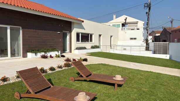 A modern house with a red-tiled roof, a covered patio with two wooden lounge chairs and a hat, and a well-maintained lawn. - Charming Aguçadoura House, 2 Min to Beach • Garden & Pet-Friendly (Aguçadoura)