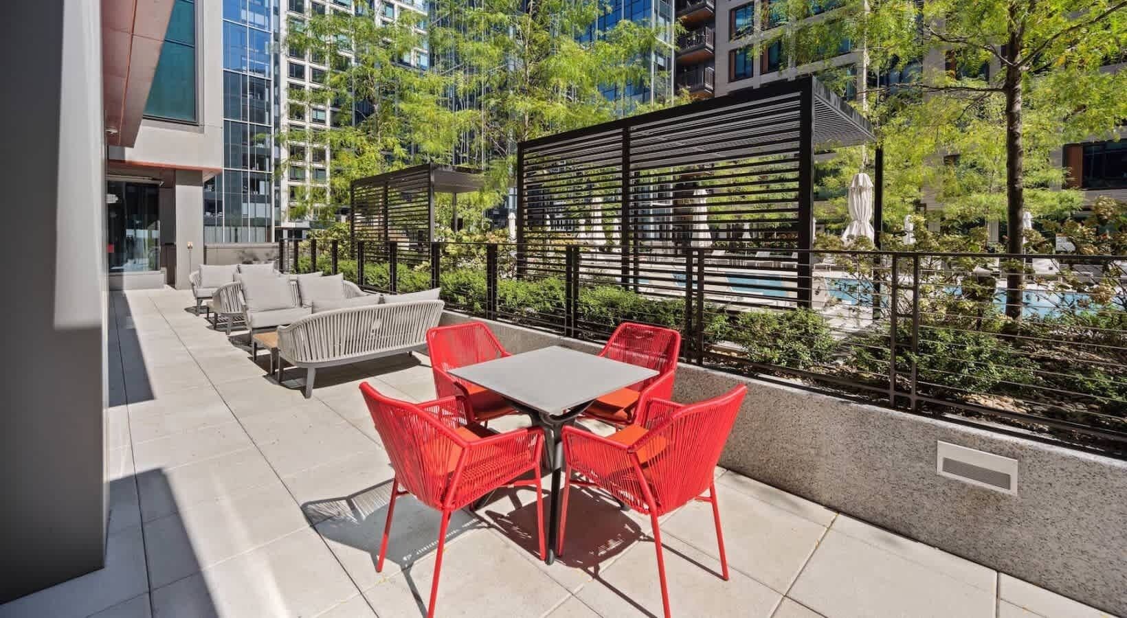 A rooftop patio with a table and chairs, surrounded by a metal fence and greenery.