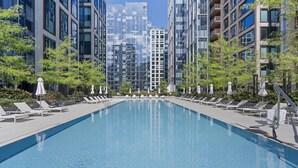 Une piscine moderne, dotée de chaises longues et de parasols, entourée d’immeubles imposants et de verdure.