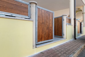 A row of closed wooden doors with metal frames, set against a yellow wall with a brick pathway. - Apartment 'Casa Dell'Oleandro' with Private Terrace, Wi-Fi and Air Conditioning (Olbia)