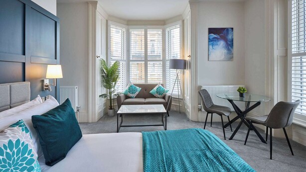 Een hotelkamer met een bed, een bank, een tafel met stoelen en uitzicht op de stad door grote ramen.