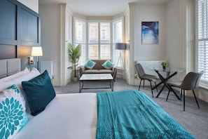 Ett hotellrum med en säng, en soffa, ett bord med stolar och utsikt över staden genom stora fönster.