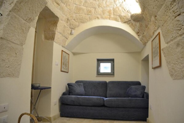 Un estrecho pasillo con paredes de piedra, un sofá gris y una pequeña mesa, que conduce a una habitación con ventana.