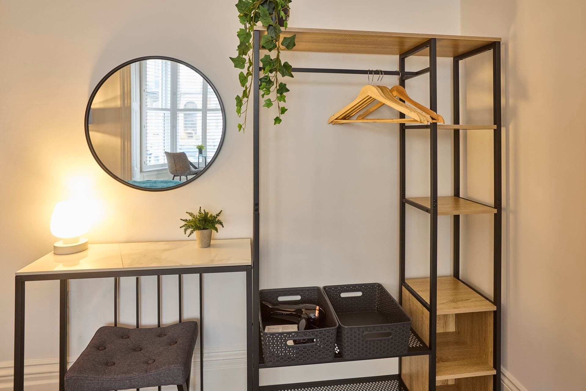 Een compacte kamer met een ronde spiegel, een klein tafeltje met een lamp, een potplant, een grijze gestoffeerde bank en een metalen rek met hangende kleding en opbergmanden.